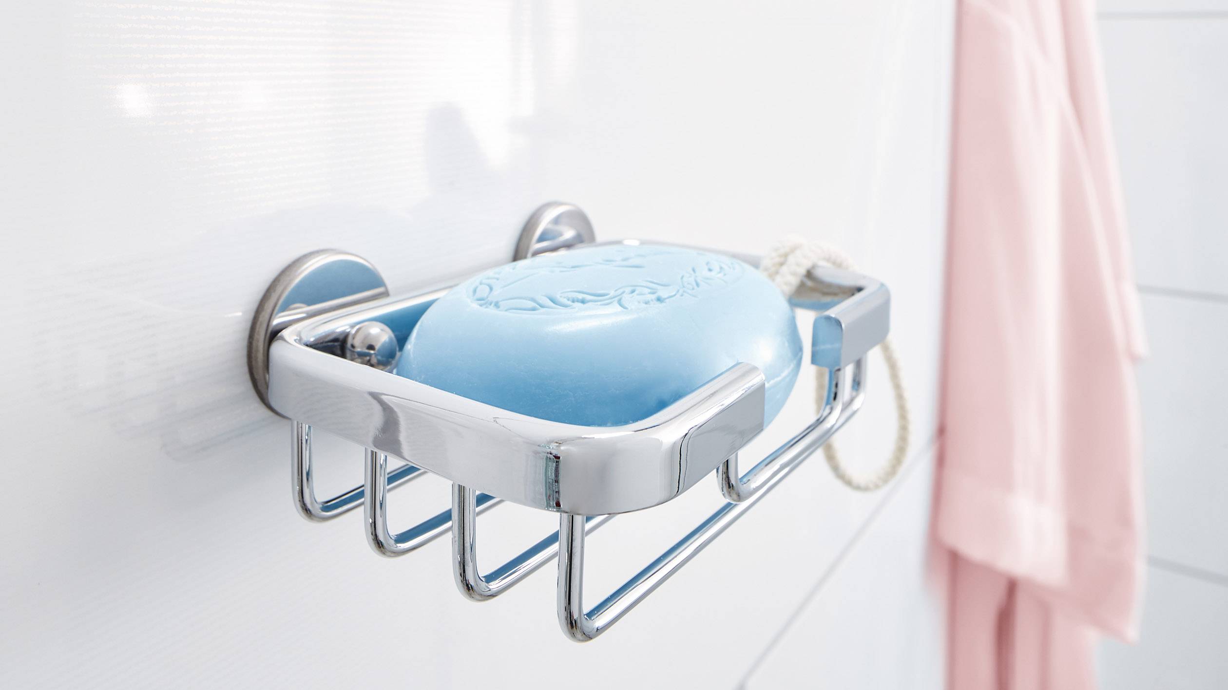 Modré mýdlo v kovovém držáku na bílé stěně, s růžovým župánem v pozadí. Koupelnové příslušenství.