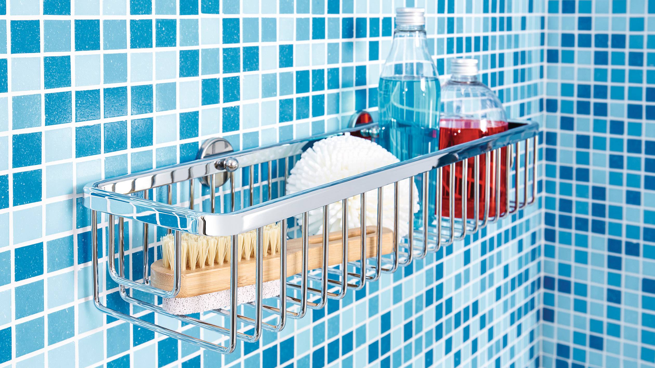 Kovová polička v sprše umístěná na modře obložené zdi, obsahující kartáč, sprchový gel a houbičku.