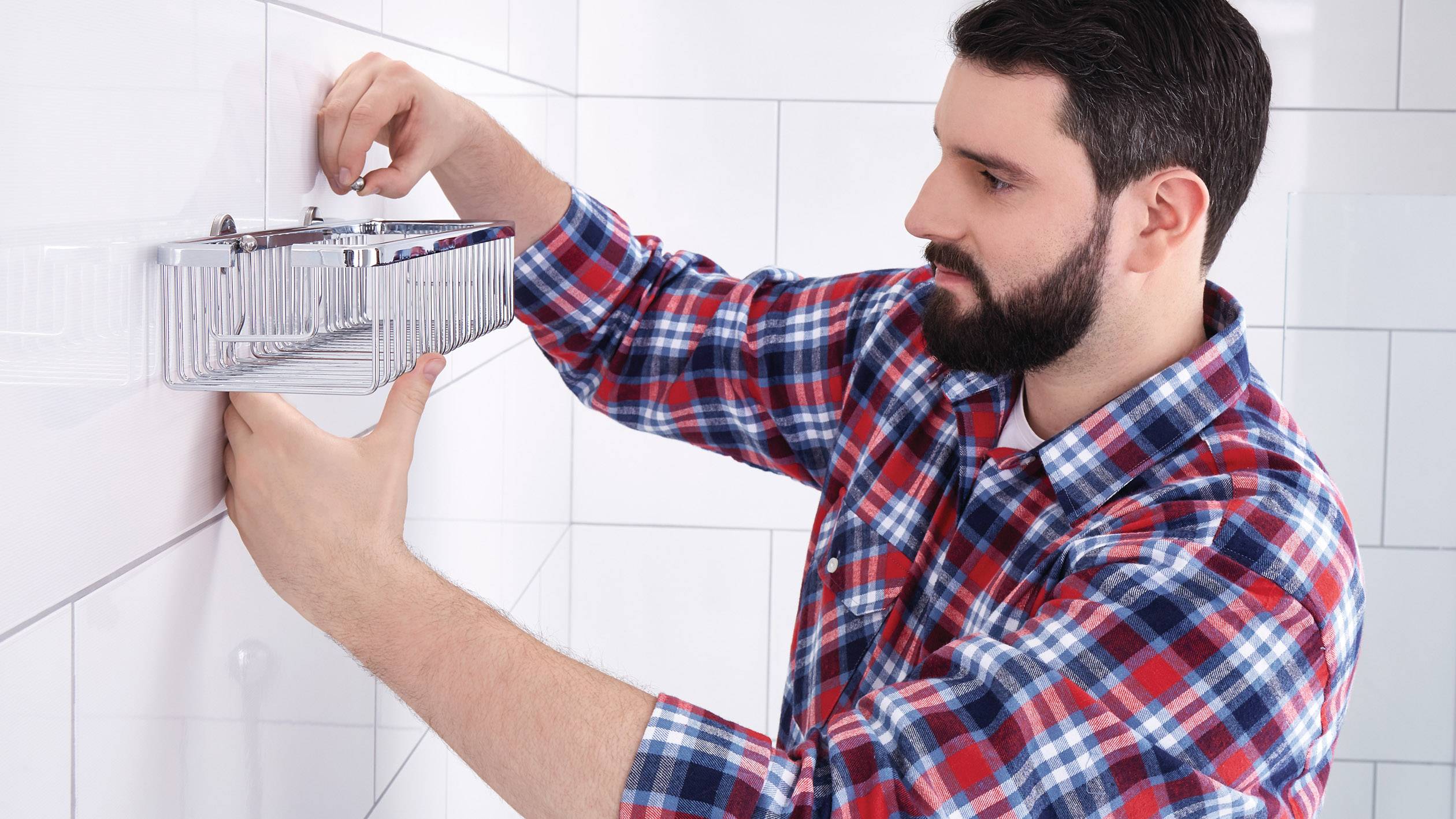 Muž montuje kovový regál na obloženou stěnu koupelny. Má na sobě kostkovanou košili a používá nářadí.