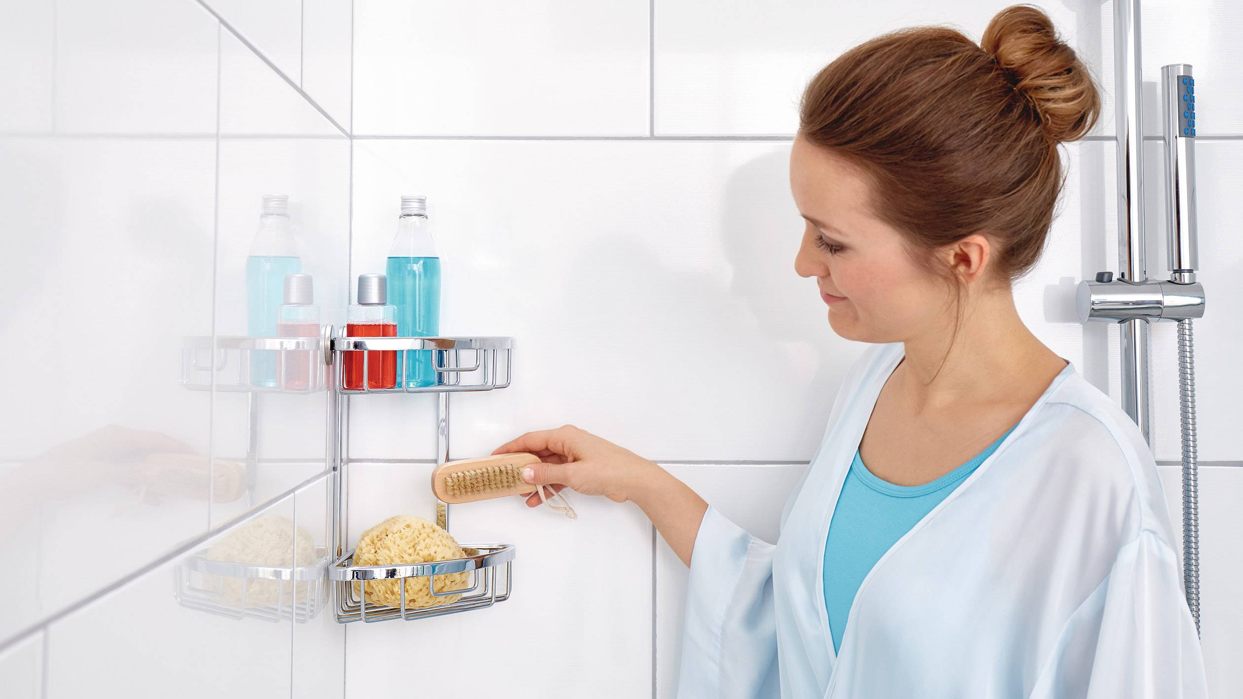 Žena stojí v koupelně, drží kartáč a dívá se na sprchové potřeby na nástěnné polici. Má na sobě světle modrý župan.