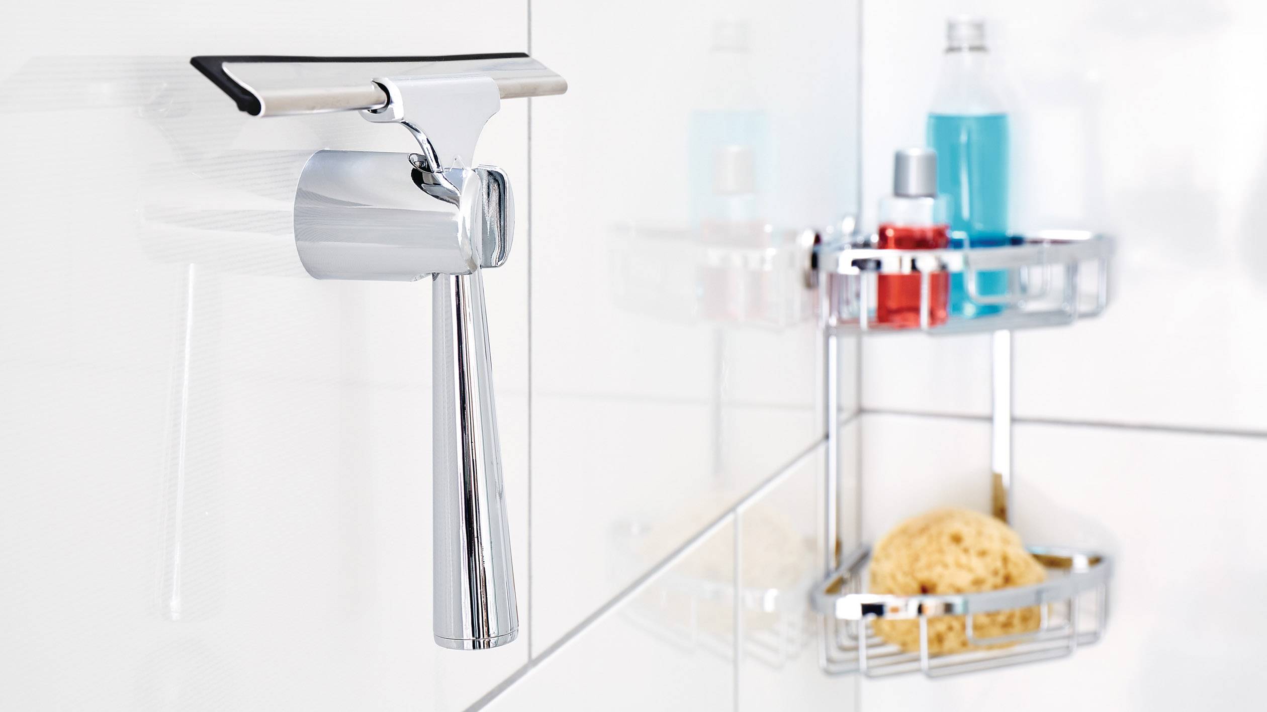 Chromové baterie na bílé koupelnové stěně, vedle police s hygienickými produkty a houbou, odrážející moderní a čistý interiér.