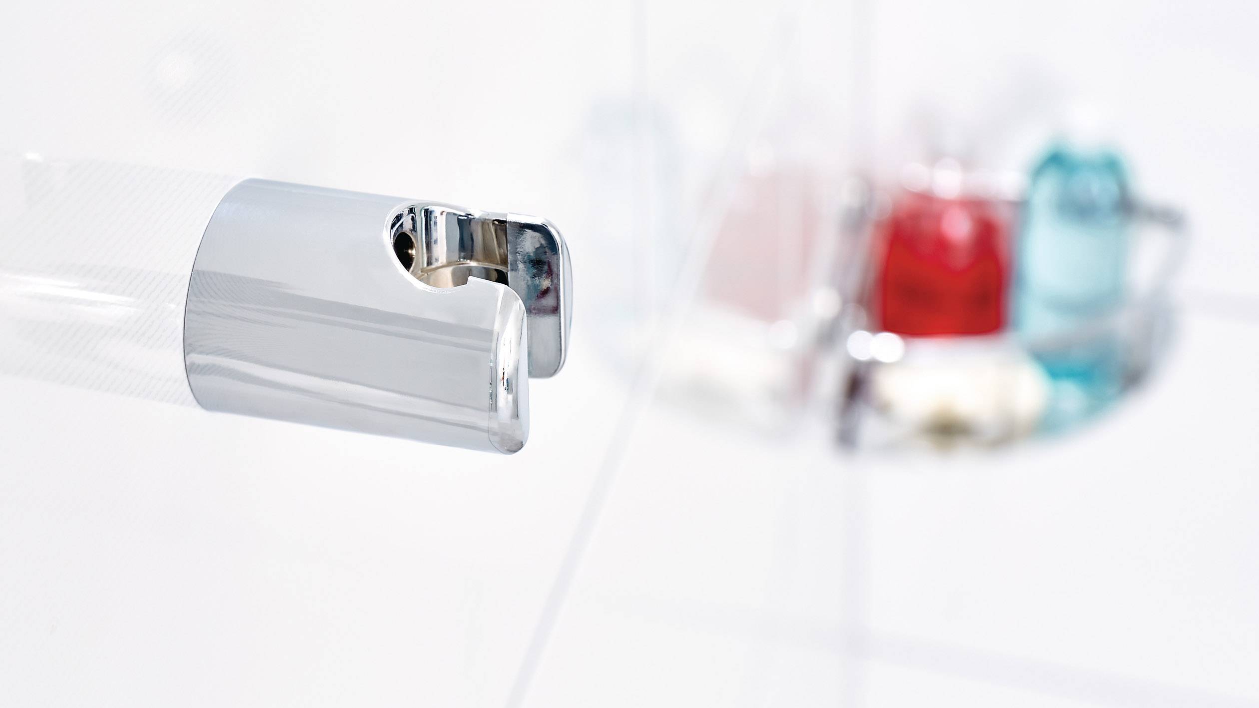 Detailní záběr lesklého držáku sprchy, v pozadí rozmazané barevné hygienické produkty.