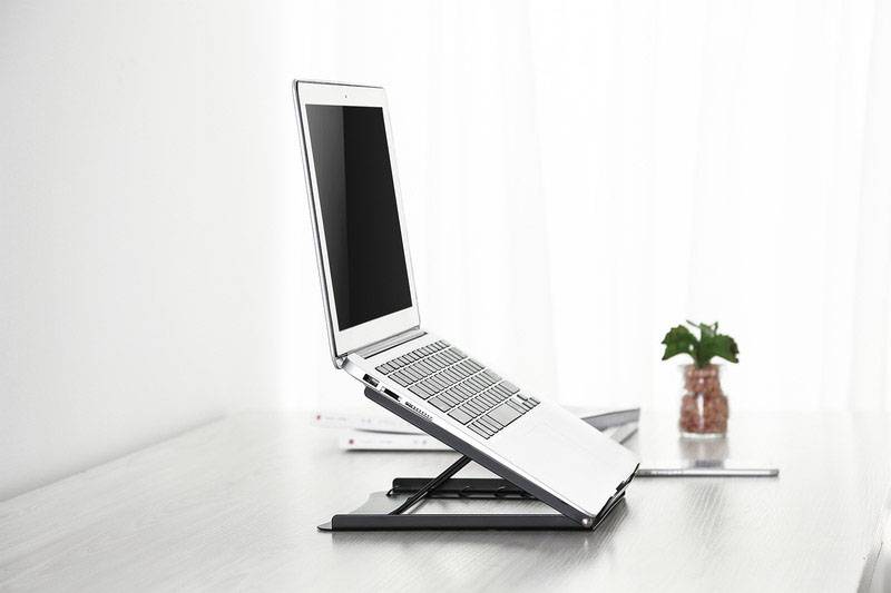 Notebook leží na nastavitelném stojanu na psacím stole, v pozadí jsou vidět knihy a malá rostlina.