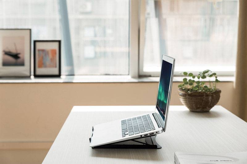 Stříbrný laptop na stole obklopený rostlinami a zarámovanými obrázky v pozadí, světlo dopadá skrze velké okno.