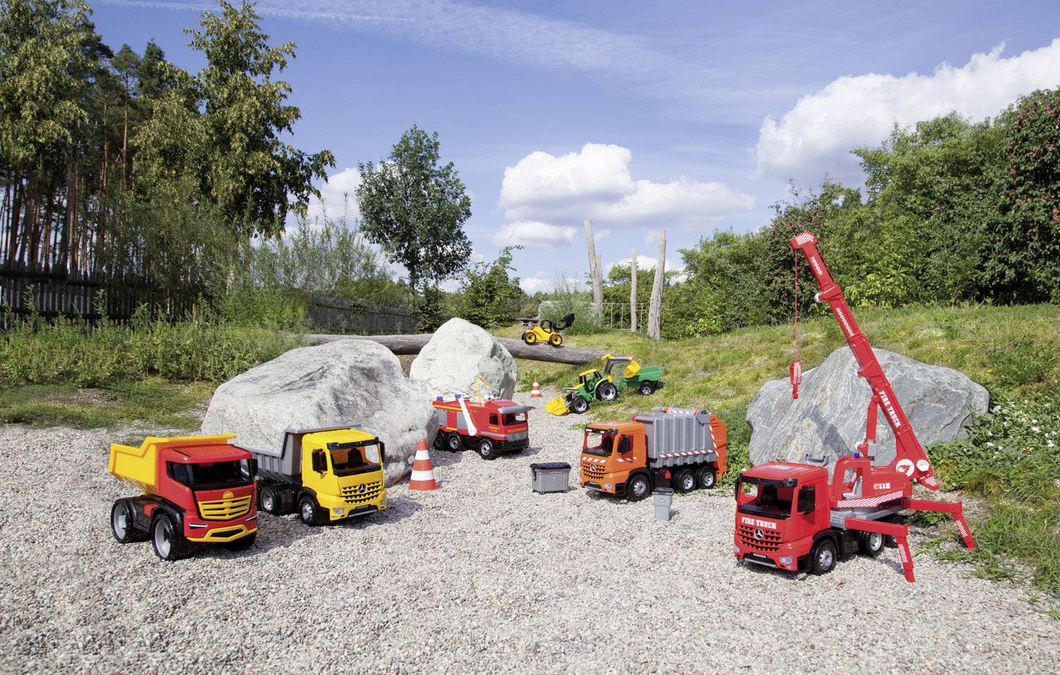 Hračkářské nákladní auto a jeřáb stojí na štěrkovém prostranství venku, obklopené stromy a skalami. Veselá venkovní hrací scéna.