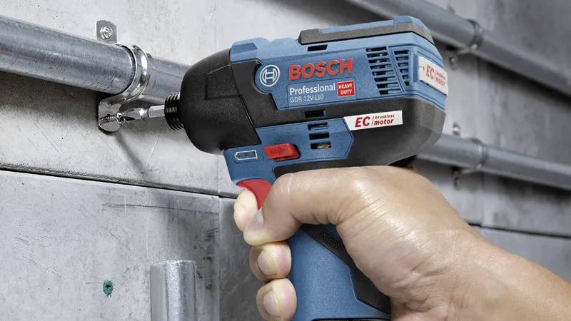 Ruka používající profesionální elektrický šroubovák Bosch pro upevnění šroubů na kovovém potrubí připevněném k betonové stěně.