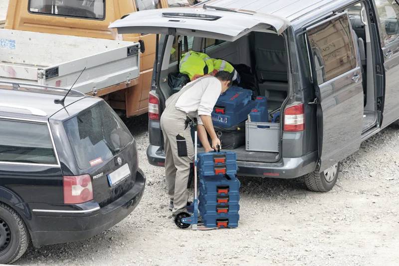 Dělník nakládá naskládané montážní kufry do šedého dodávkového vozu na štěrkovém povrchu s parkujícími vozidly kolem.