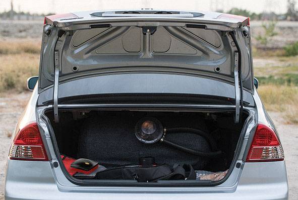 Auto s otevřeným zavazadlovým prostorem, které obsahuje plynovou láhev a nářadí. V pozadí je venkovská silnice.