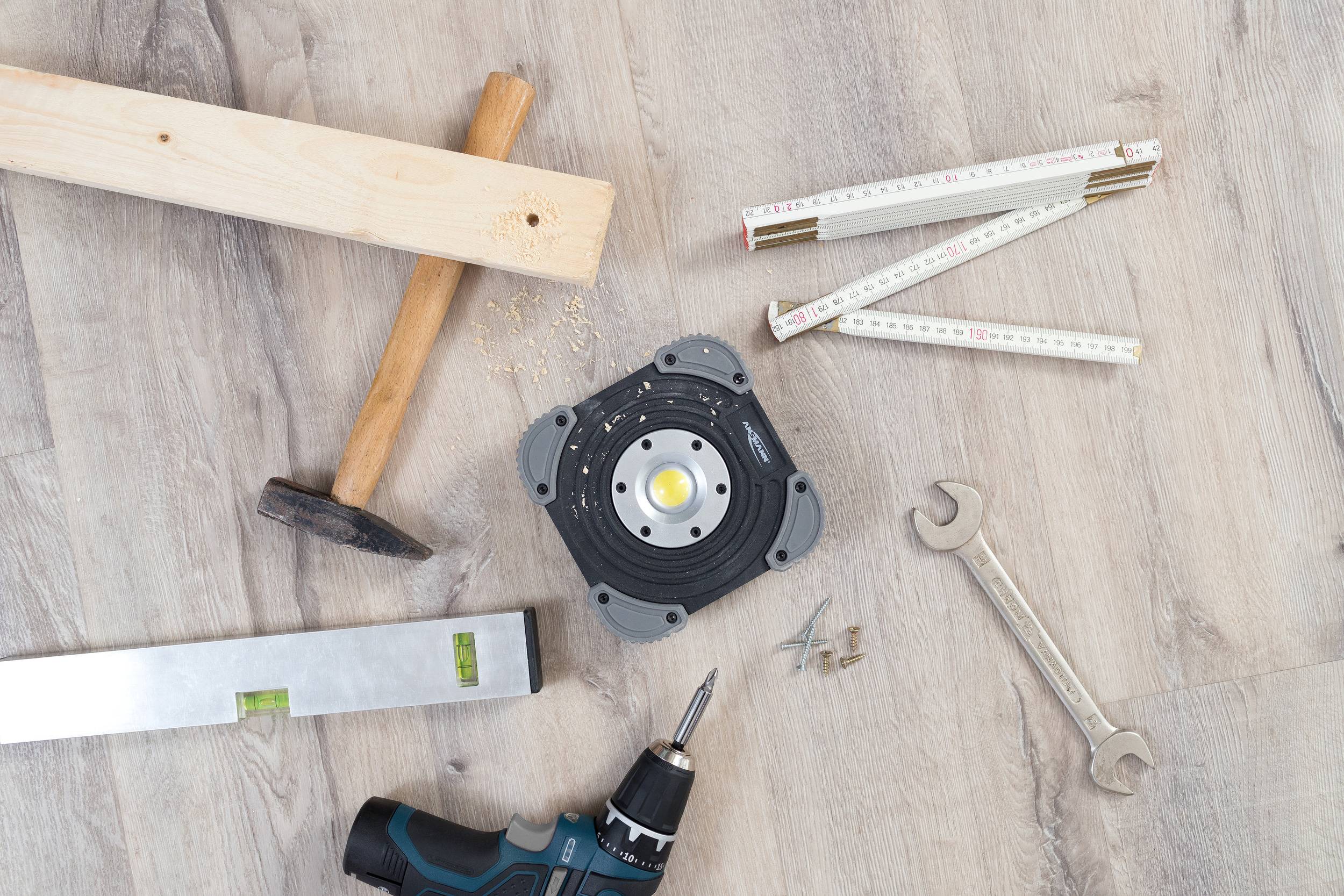 'Stavební nářadí na dřevěné podlaze: dřevěný špalík, kladivo, metr, klíč, vodováha, vrtačka a šrouby.'