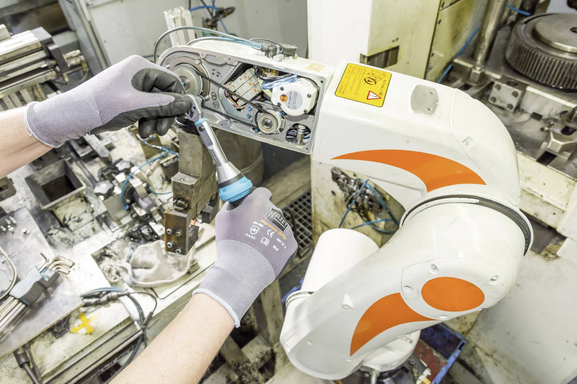 Dělník montuje nástroj na rameno robota v prostředí dílny. Robot má bílé a oranžové detaily.