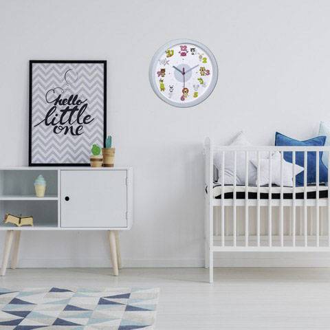 Dětský pokoj s bílou postýlkou, nástěnnými hodinami a policí s dekoracemi. Na stěně visí obrázek s nápisem 'Hello little one'.
