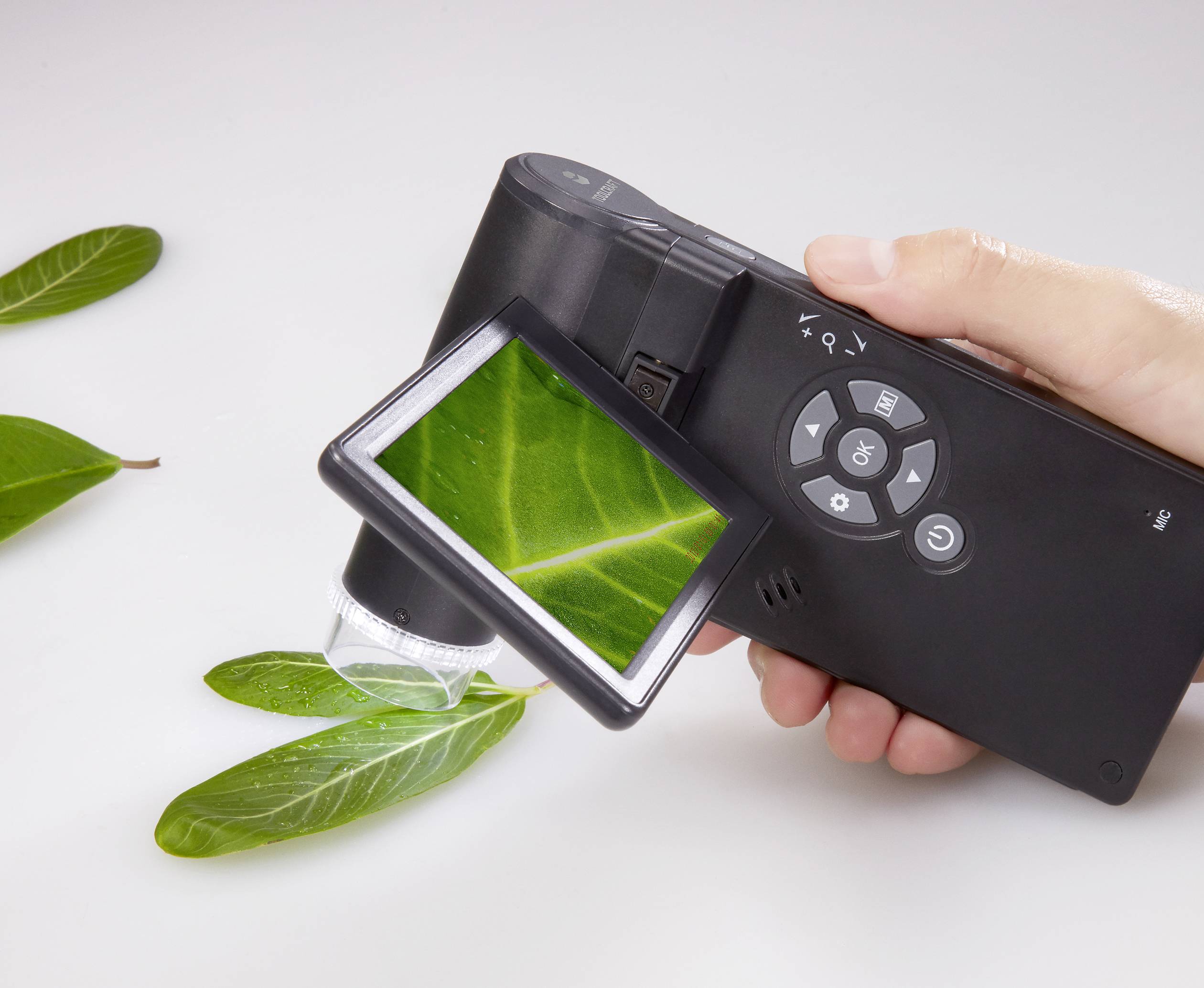 Detailní záběr ruky držící digitální mikroskop, který zobrazuje zvětšený obraz zeleného listu na displeji.