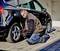 Osoba klečí na zemi a používá nástroj ke kontrole nebo opravě pneumatik černého automobilu.