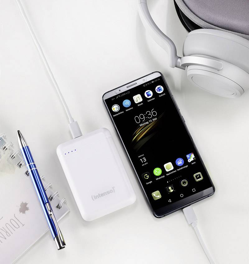 Smartphone s černou obrazovkou zobrazuje 09:36. Je připojeno k bílé powerbance a obklopeno sluchátky, perem a poznámkovým blokem.