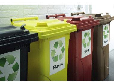 Pět odpadkových nádob s recyklačními symboly, označených pro plast, sklo a papír před bílou stěnou.