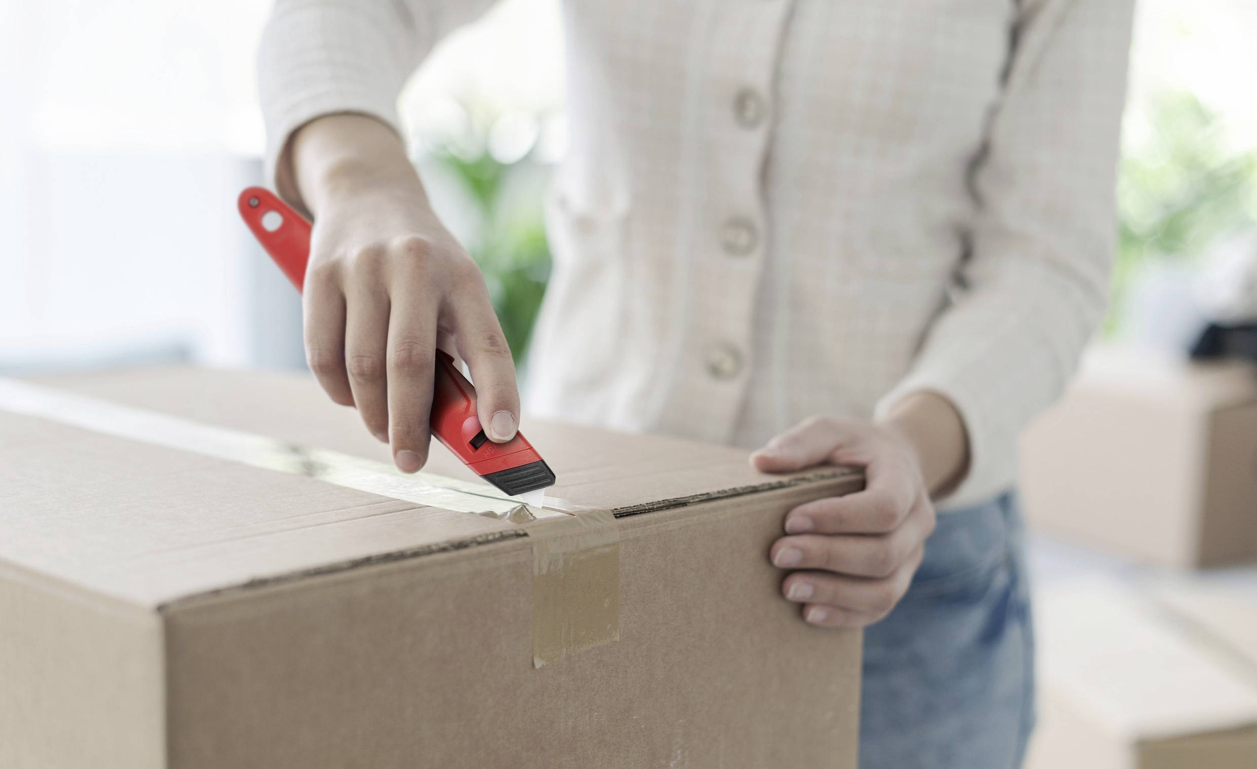 Osoba otevírá velkou krabici červeným řezákem. V pozadí jsou rozmazaně vidět zabalené bedny.