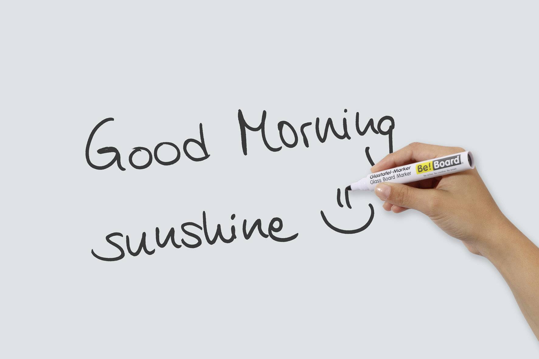 Ruka píše 'Good Morning sunshine' černým perem na bílou tabuli, doprovázenou úsměvem.