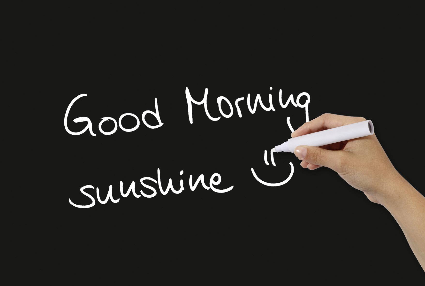 Ruka píše bílou tužkou na černou tabuli: 'Good Morning sunshine', doplněno úsměvem.
