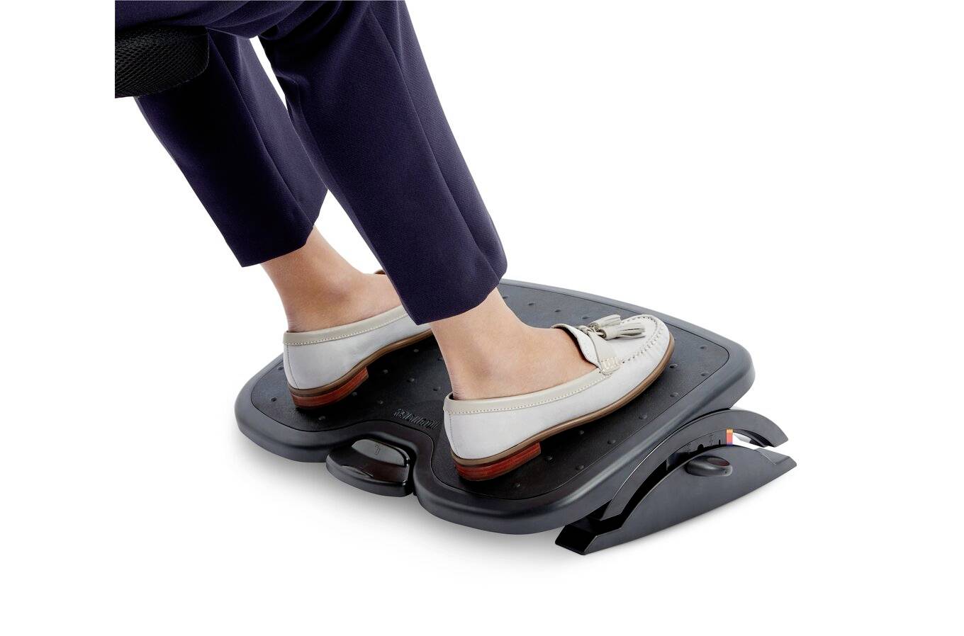 Nohy člověka v třásňových mokasínách spočívají na černém ergonomickém podnožníku, což zdůrazňuje pohodlí a podporu správného držení těla v kancelářském prostředí.