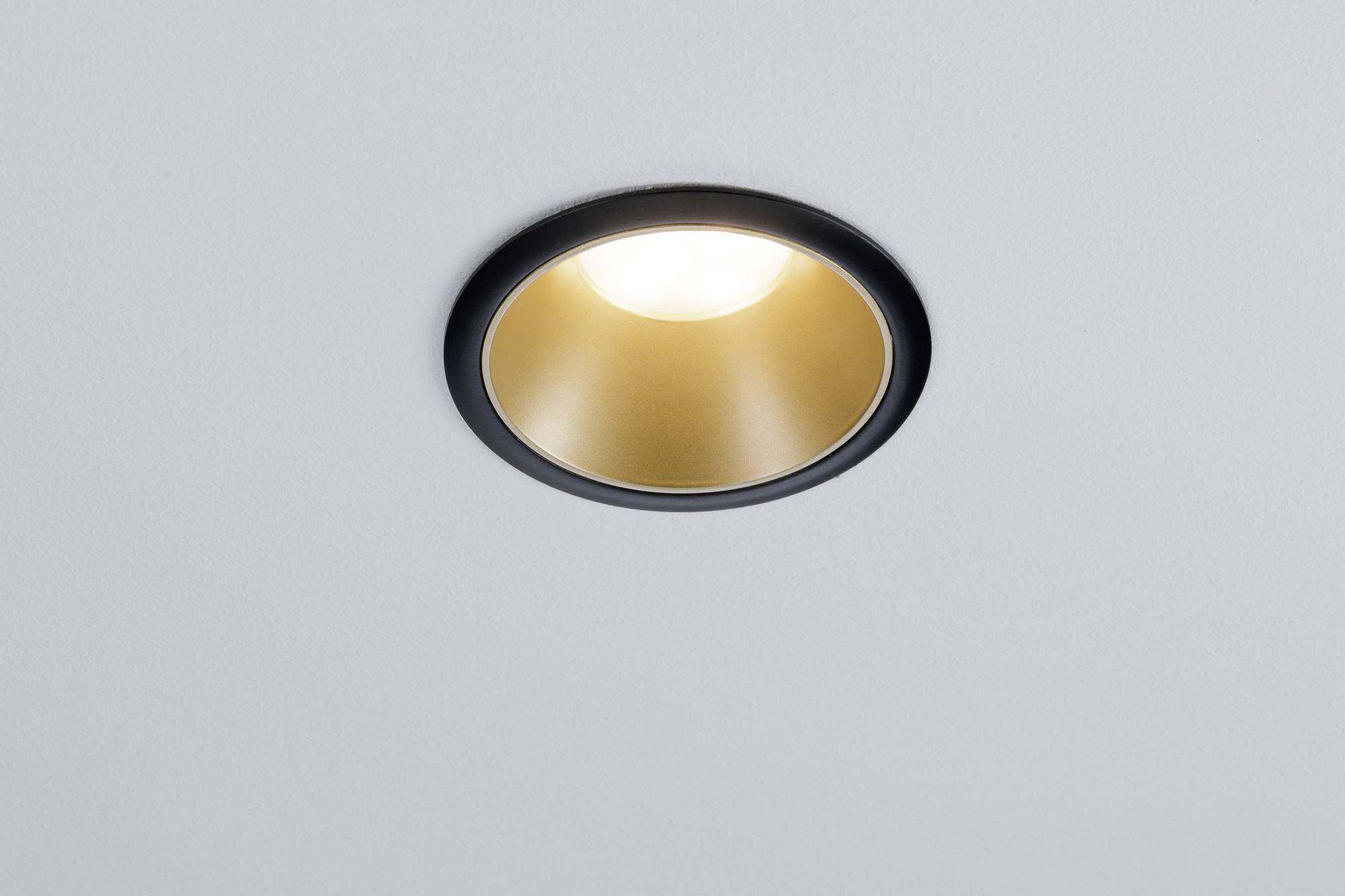 Stropní lampa s černým rámem a zlatým vnitřkem vyzařuje teplé světlo z bílého stropu.