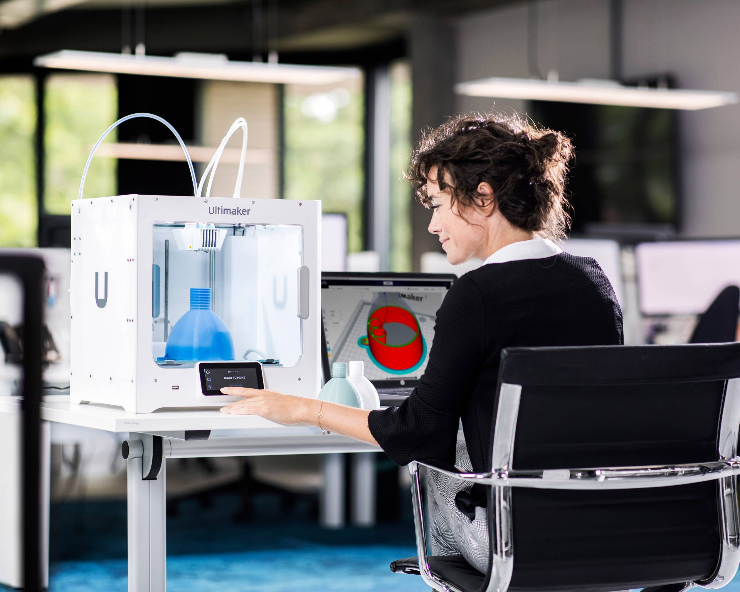 Osoba sedí u psacího stolu a obsluhuje 3D tiskárnu, přičemž na počítačové obrazovce je viditelná grafika.