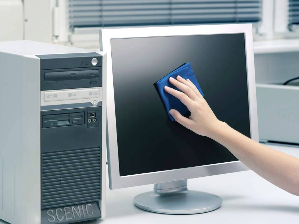 Osoba čistí monitor počítače s plochou obrazovkou modrým hadříkem v kancelářském prostředí, vedle stolního počítače.