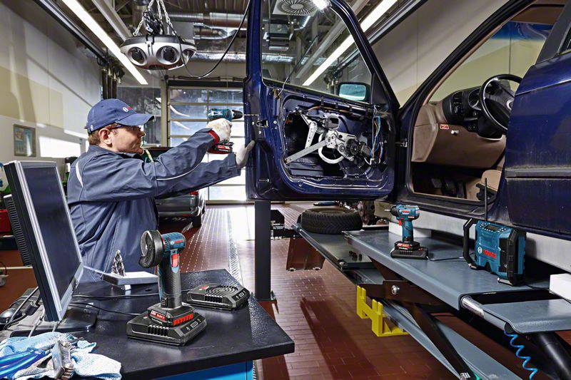 Mechanik opravuje řidičovy dveře nadzvednutého automobilu v autodílně. Různé nástroje jsou rozložené na stole.