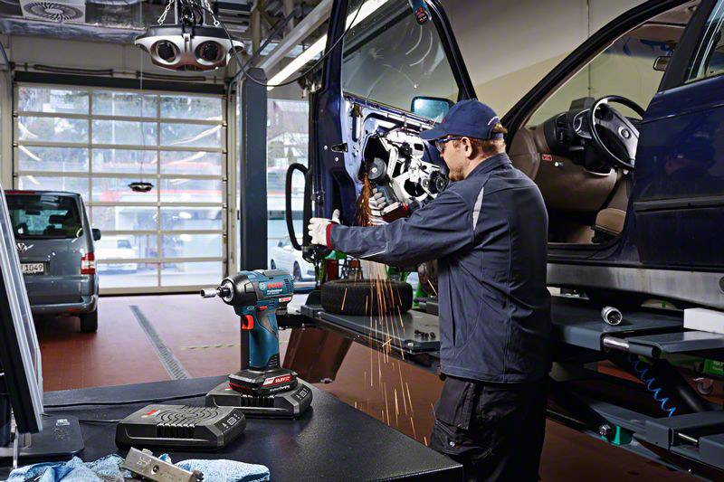 Mechanik opracovává dveře vozidla nástrojem v autoservisu. Od kovu odletují jiskry. V pozadí stojí další automobily.