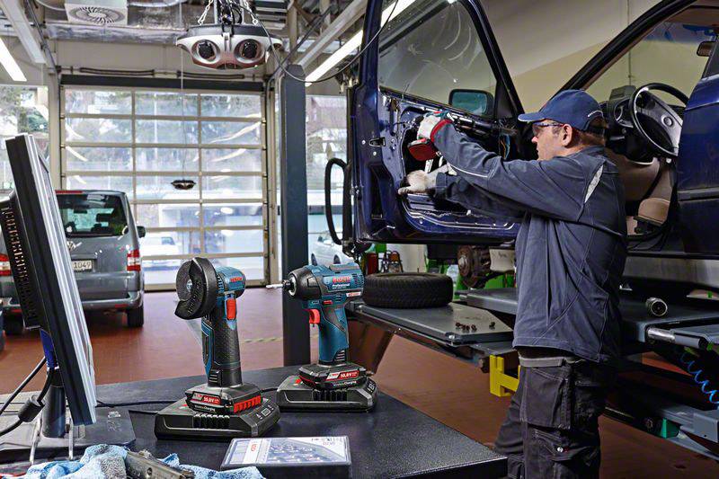 Mechanik v autodílně opravuje palubní desku automobilu. Dva akumulátorové šroubováky leží v popředí na stole.