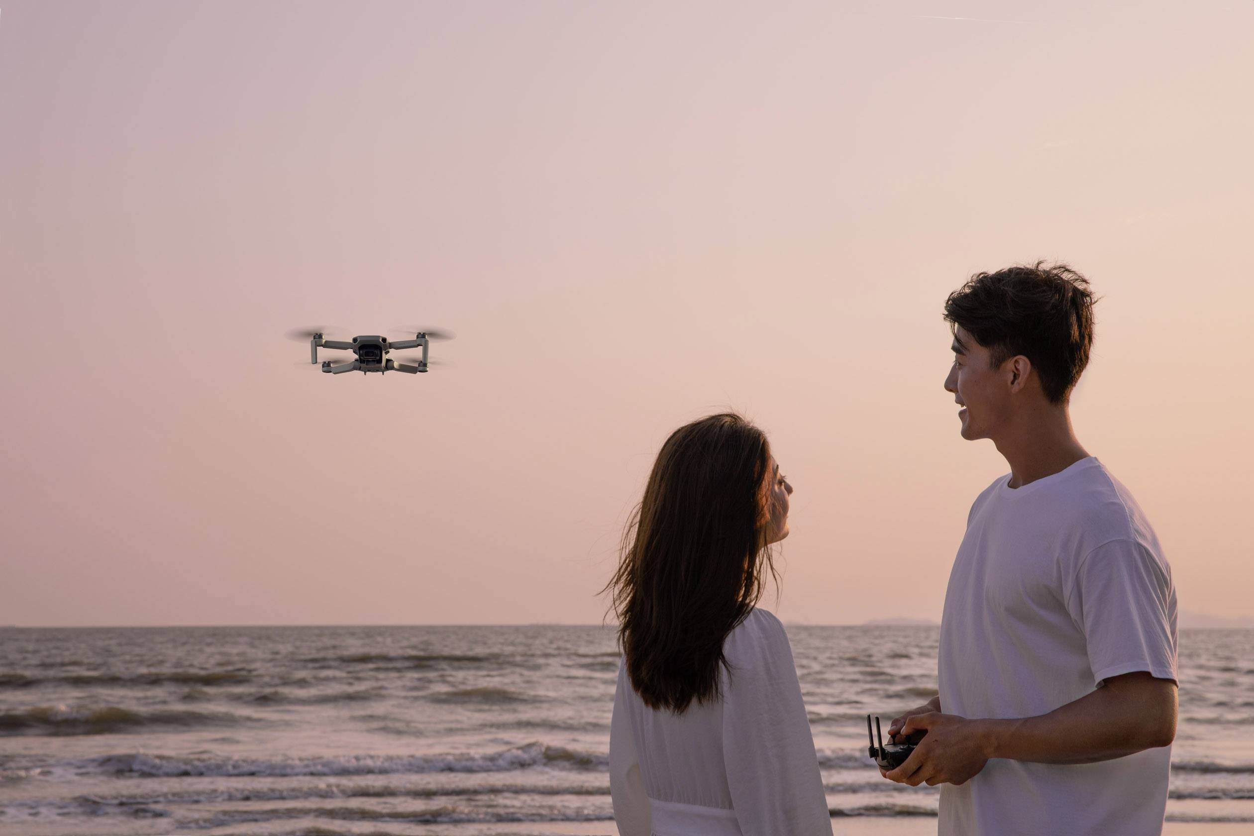 Muž a žena sledují při západu slunce létající dron nad mořským horizontem.