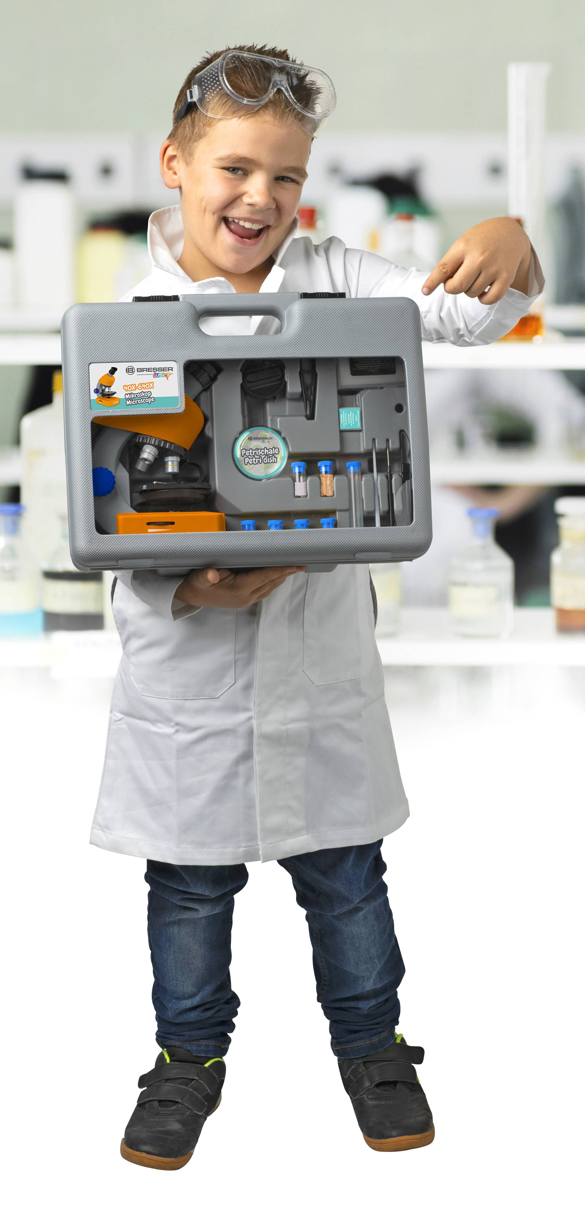 Dítě v laboratorním plášti a ochranných brýlích hrdě drží kufřík s různými laboratorními pomůckami v laboratorním prostředí.