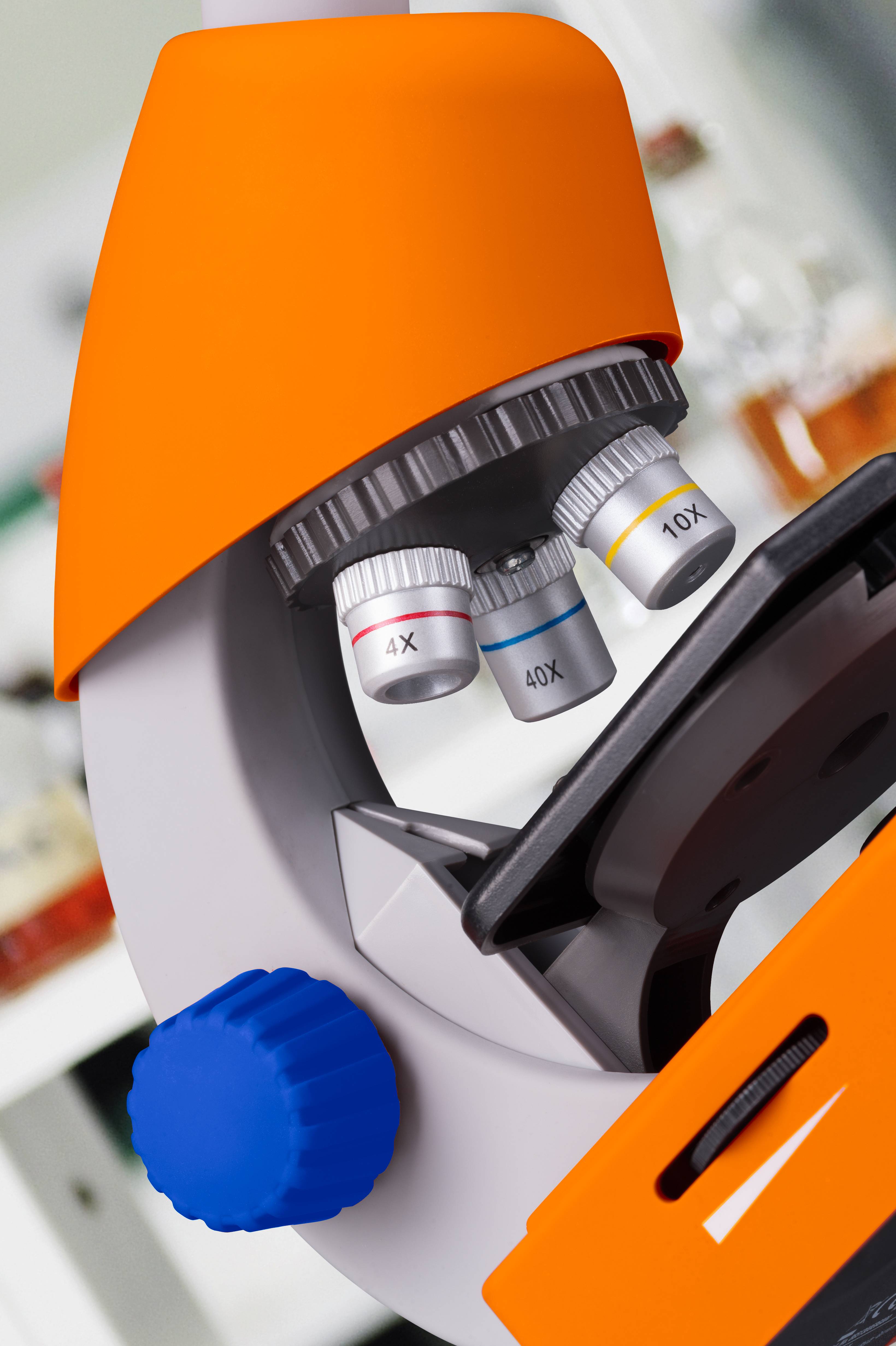 Mikroskop se čtyřmi objektivy a oranžovo-modrým barevným designem, v pozadí rozmazané laboratorní vybavení.