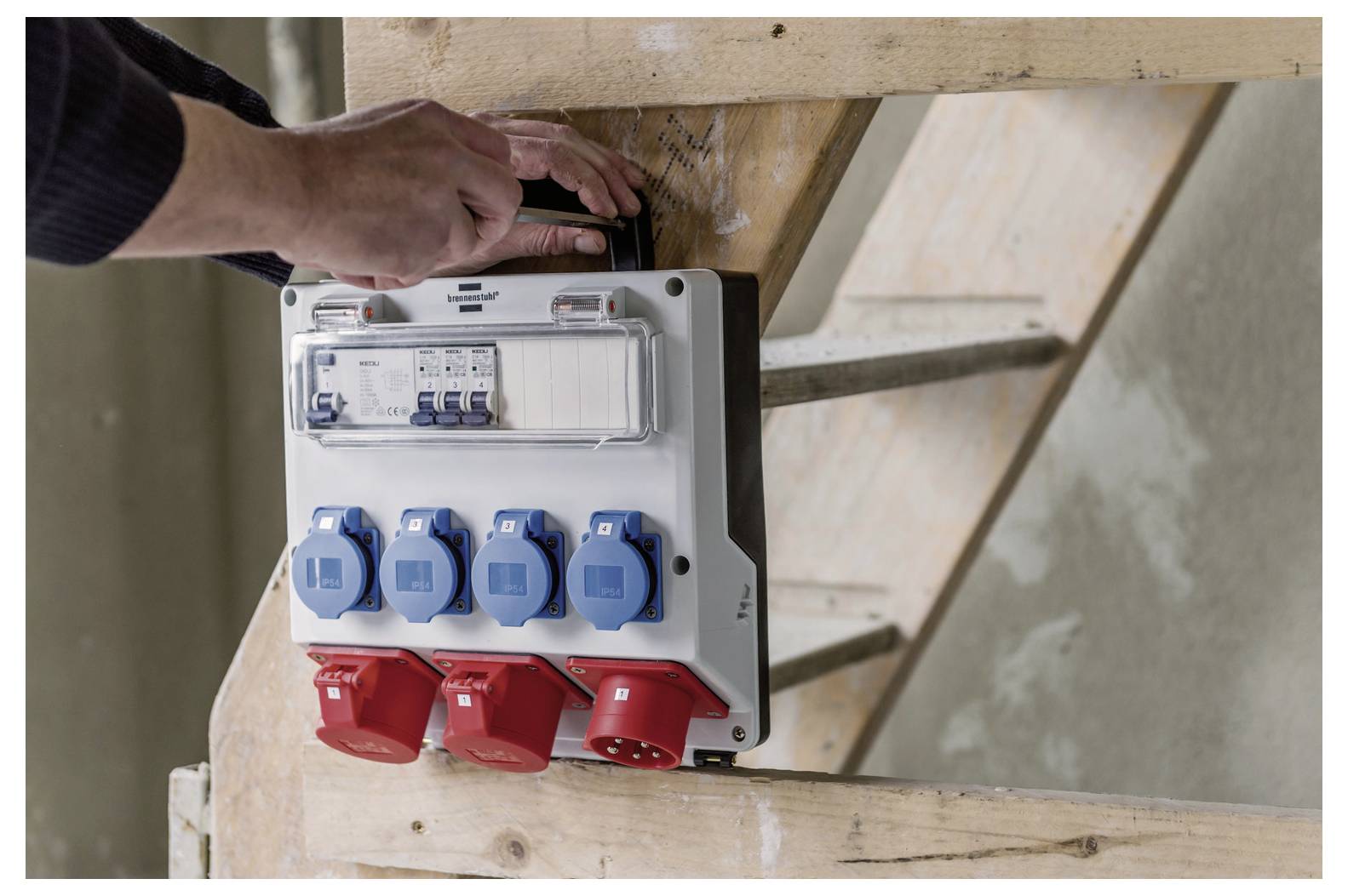 Ruka instaluje elektrickou rozvodnou skříň s modrými a červenými průmyslovými zásuvkami namontovanými na dřevěném lešení.