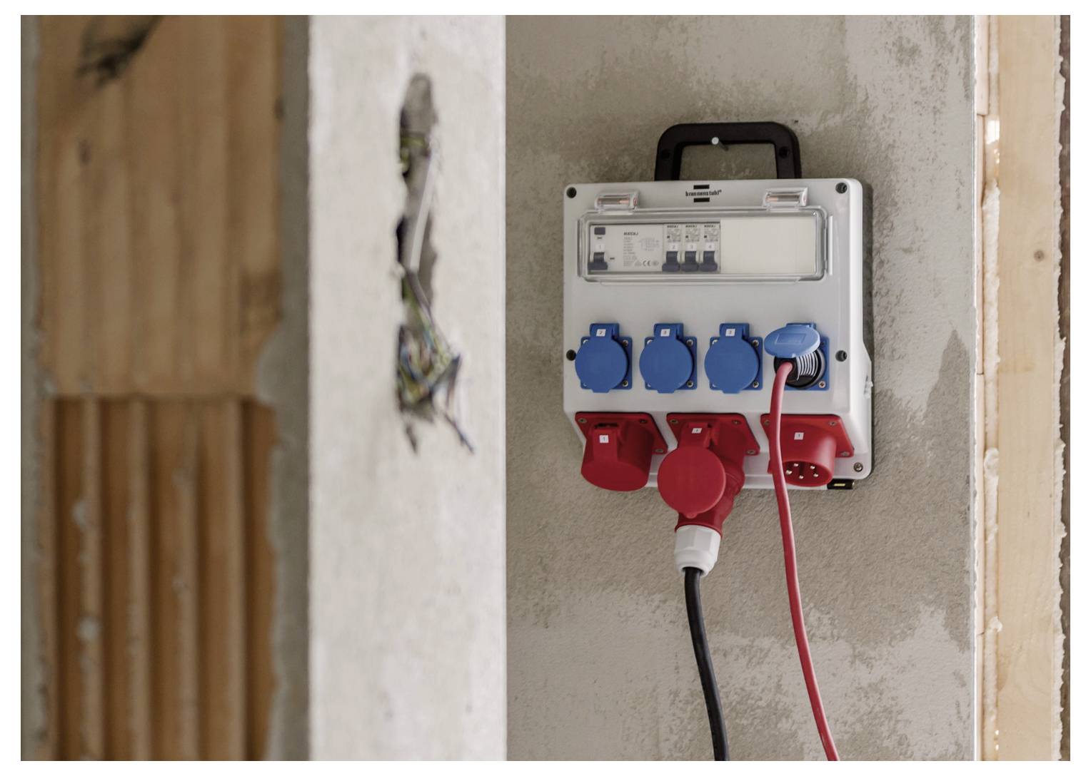 Nástěnná elektrická rozvodnice s více červenými a modrými zásuvkami napájení a zastrčenými kabely v nedokončené vnitřní stěně.