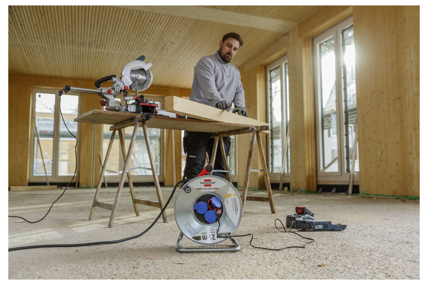 Muž používá pokosovou pilu na řezání dřevěné desky v domě ve výstavbě, s bubnem elektrického kabelu a nástroji na podlaze.