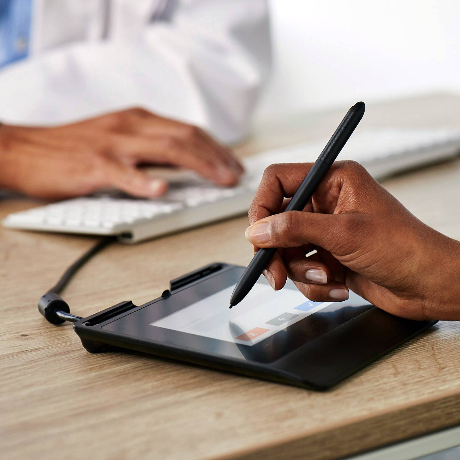 Osoba podepisuje dokument na digitálním tabletu stylusem, zatímco druhá osoba používá klávesnici.
