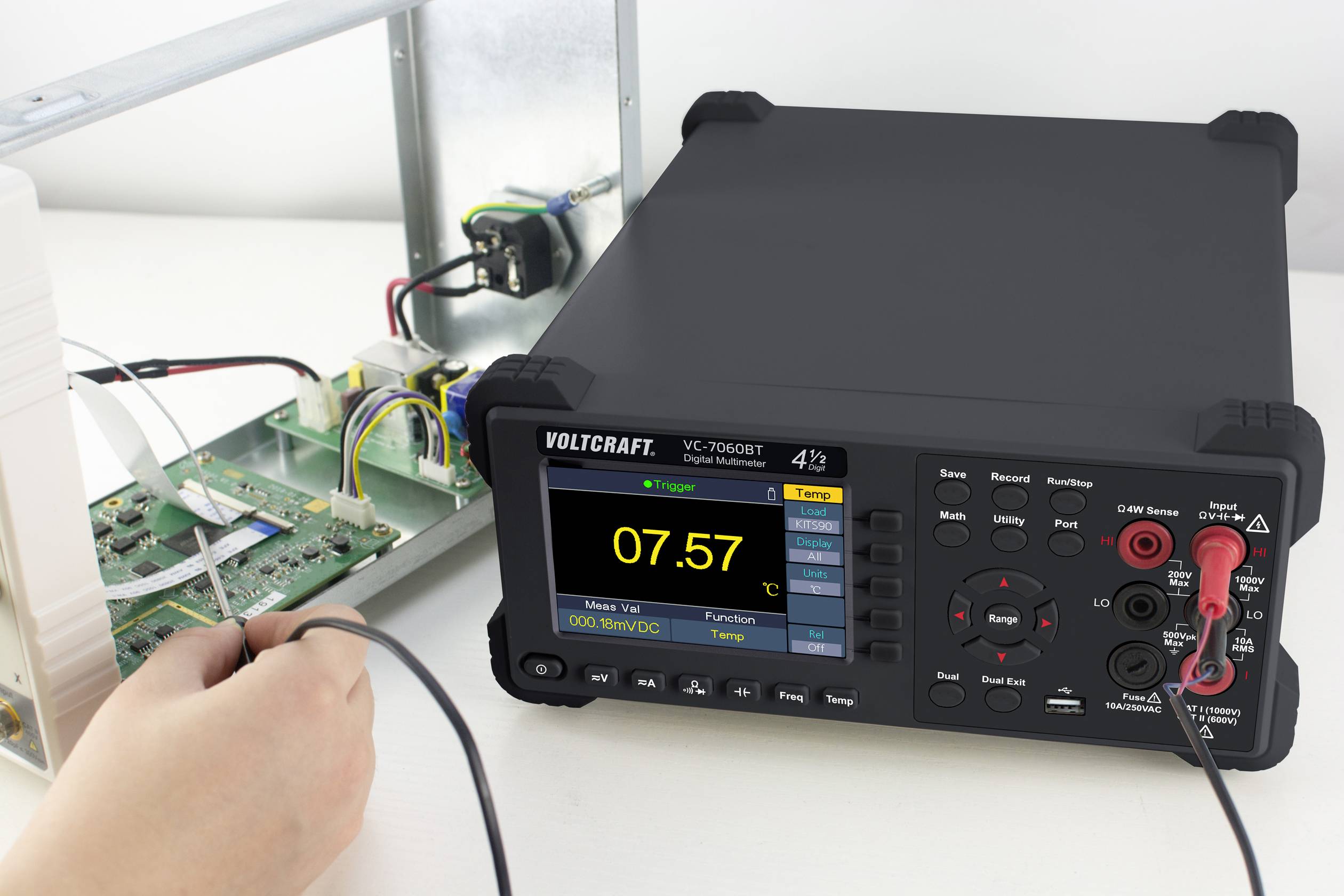 Osoba měří elektrické napětí digitálním multimetrem, který ukazuje napětí 7,57 voltů. V pozadí jsou vidět součástky elektronického obvodu.