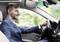 Muž v obleku řídí auto a mluví pomocí náhlavní soupravy. V pozadí jsou rozmazané zelené a bílé barvy.