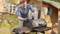 Muž s brýlemi a rukavicemi otevírá gril na zahradě. V pozadí je vidět dřevěná chata.