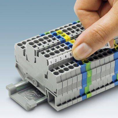 Jedna ruka zasunuje očíslovanou zástrčku do šedého, vícebarevně označeného elektrického svorkovnice.