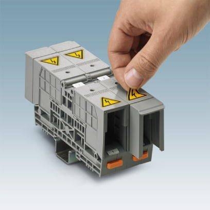 Ruka montuje šedou elektrickou svorku na kolejnici. Na svorce jsou viditelné žluté výstražné symboly s bleskem.