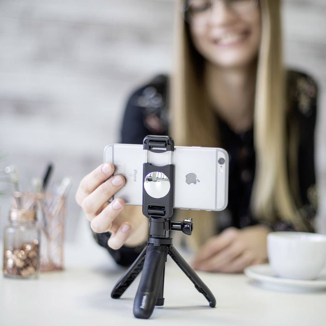 Usměvavá osoba se fotografuje smartphonem umístěným na stativu. V pozadí jsou rozmazané kancelářské potřeby.