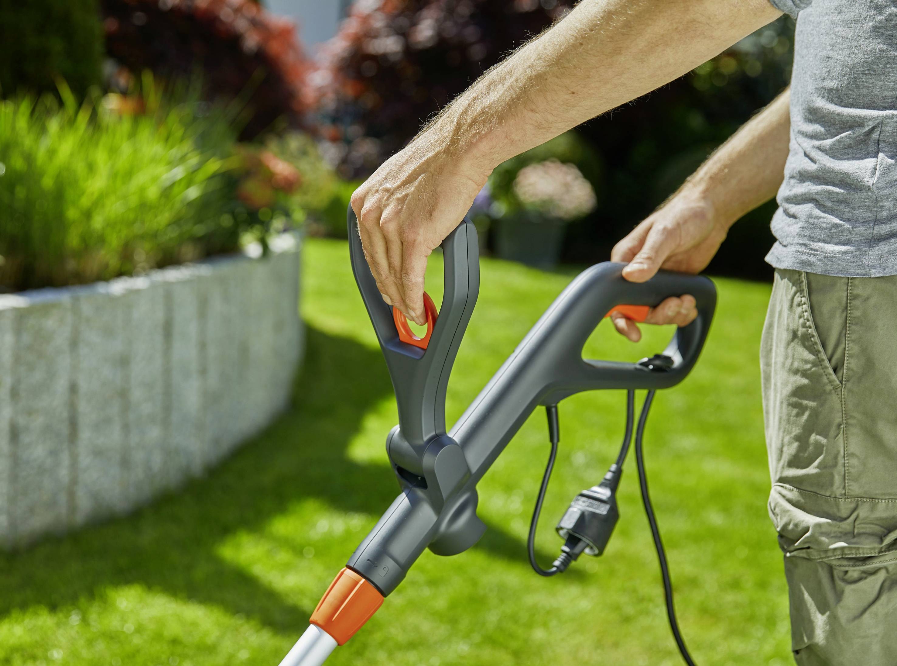 Osoba používá elektrický vyžínač k sestřihávání trávy na sluncem zalité zahradě.