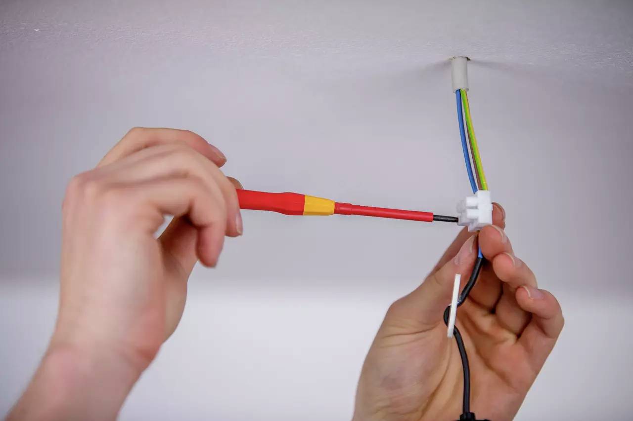 Několik rukou instaluje elektrický kabel na stropě. Šroubovák se používá k zajištění připojení.