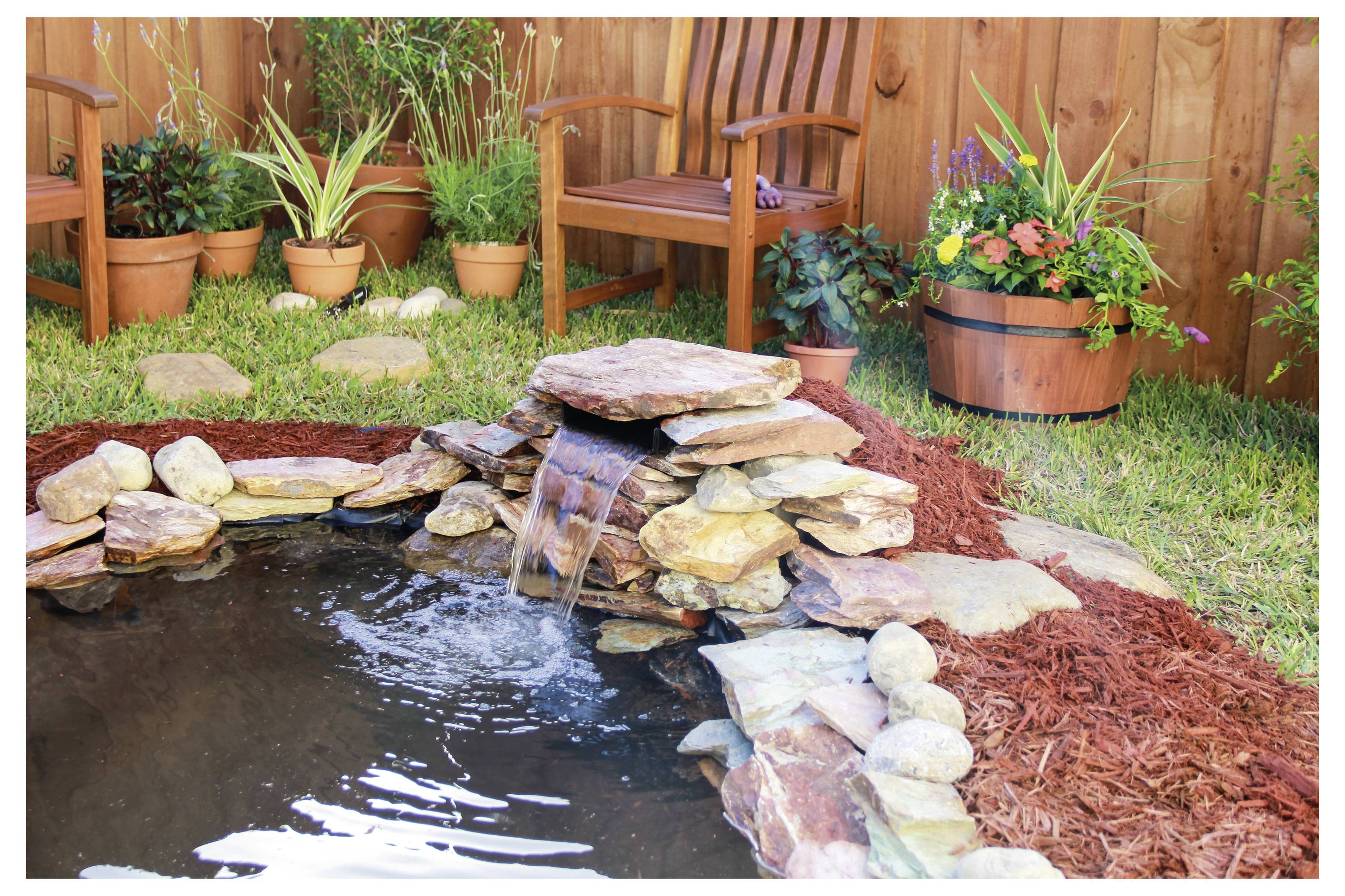 Malý zahradní rybníček s kamenným vodopádem, obklopený mulčovací kůrou a květináči s rostlinami. Poblíž jsou dřevěné židle a dřevěný sud na pěstování rostlin.