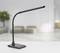 Černá LED stolní lampa s flexibilním ramenem stojí na bílém psacím stole v moderně a minimalisticky zařízené místnosti.