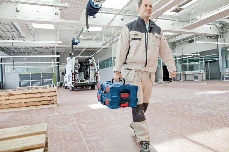 Řemeslník v pracovním oblečení nese kufřík s nářadím ve velké dílně. V pozadí stojí otevřený dodávkový vůz.