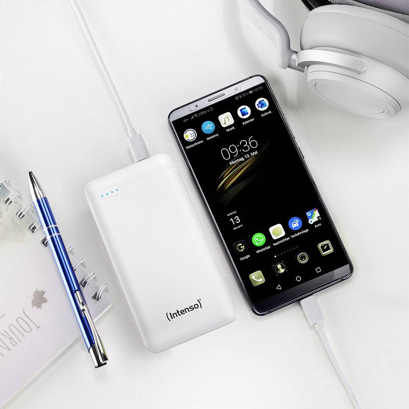Powerbanka a smartphone s indikátorem nabíjení, propojené kabelem na psacím stole s poznámkovým blokem, perem a sluchátky.