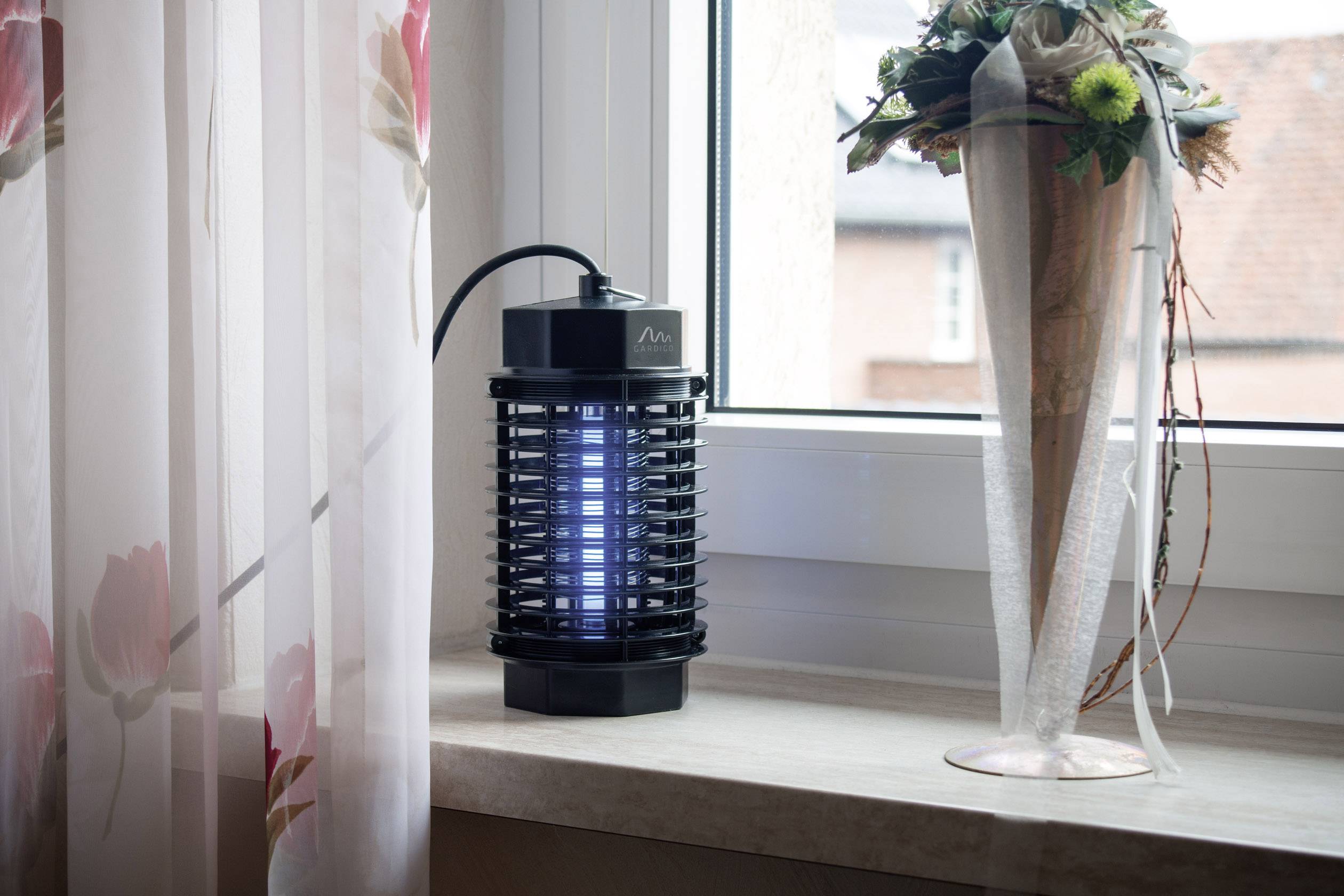 Elektrický lapač hmyzu stojí na okenním parapetu vedle vázy s květinami. Venku je vidět střecha pokrytá taškami.