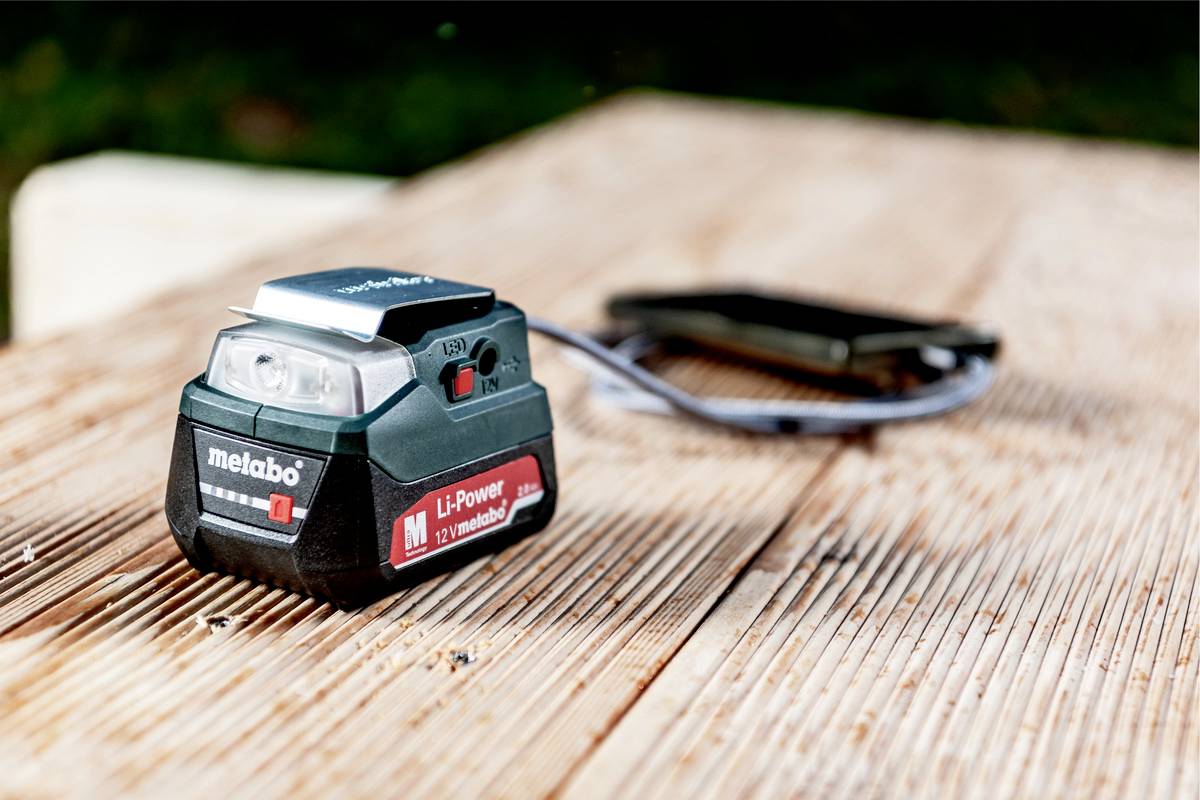 Na dřevěném stole leží Metabo Li-Power akumulátor s 12V, v pozadí rozmazaný objekt.